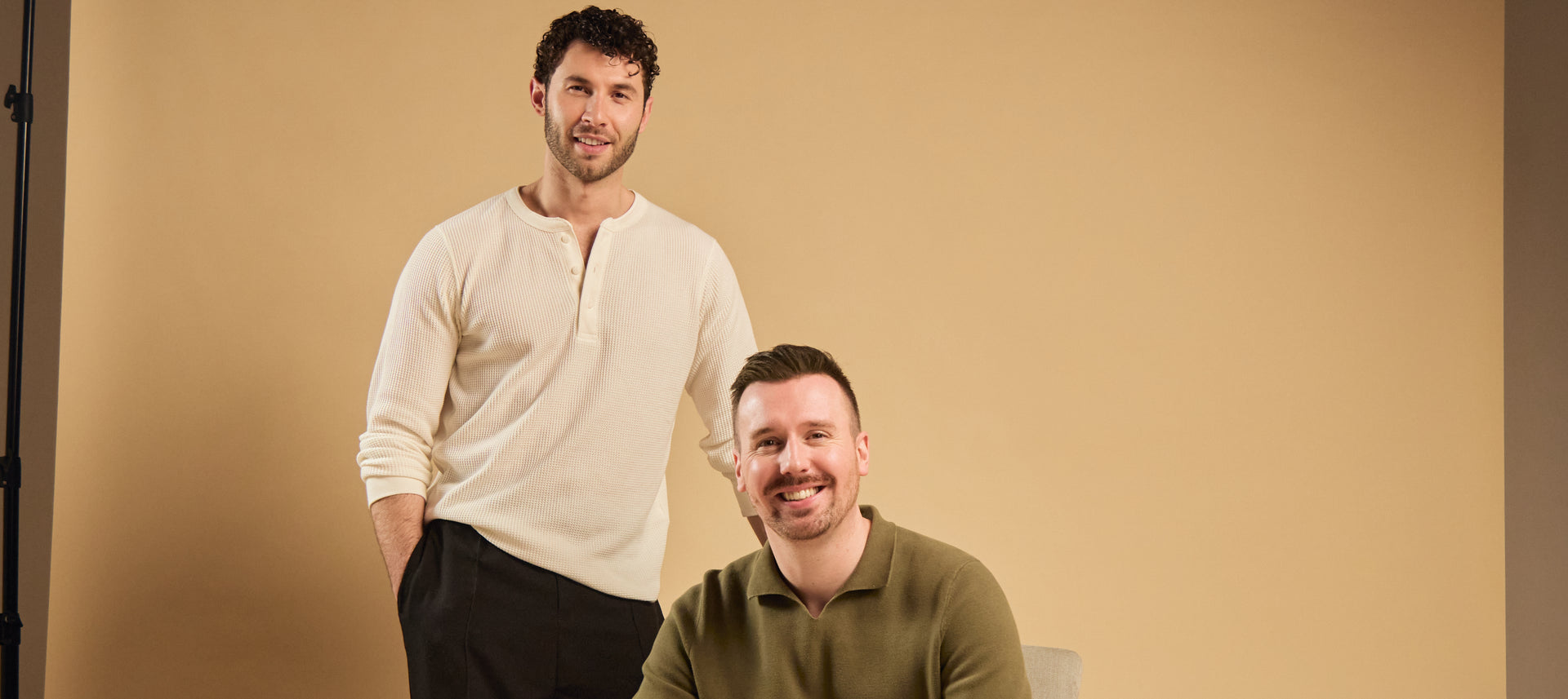 Two smiling men pose against a beige background, one standing and one seated.