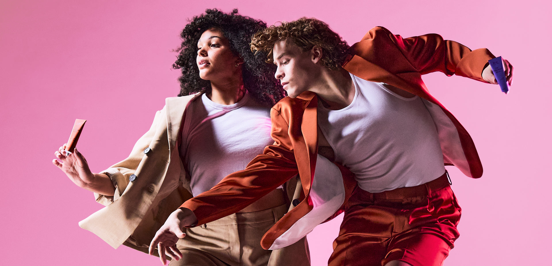 Two people in motion holding products against a pink background.